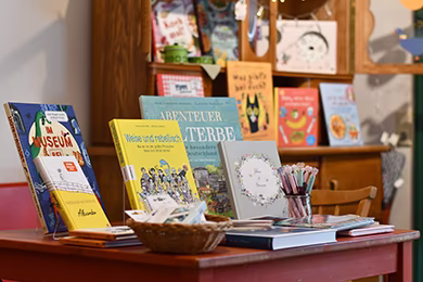 Bücher stehen auf einem roten Tisch, im Hintergrund ist unscharf eine Holzkommode mit Büchern darauf