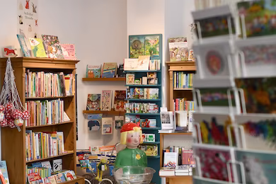 Blick in die Buchhandlung mit Postkartenständer im Vordergrund und Bücherschrank und Pixi-Figur im Hintergrund